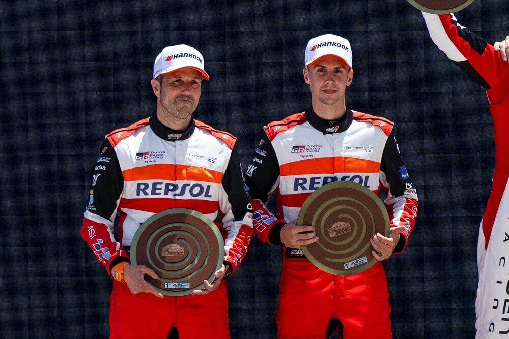 Dos pilotos de carreras posan juntos con uniformes rojos y blancos de Toyota Racing España. Sus trajes muestran el logotipo de Repsol en el centro y el de Hankook en sus gorras. Ambos sostienen placas circulares de premio con el logotipo del WRC (World Rally Championship) en la parte inferior.
