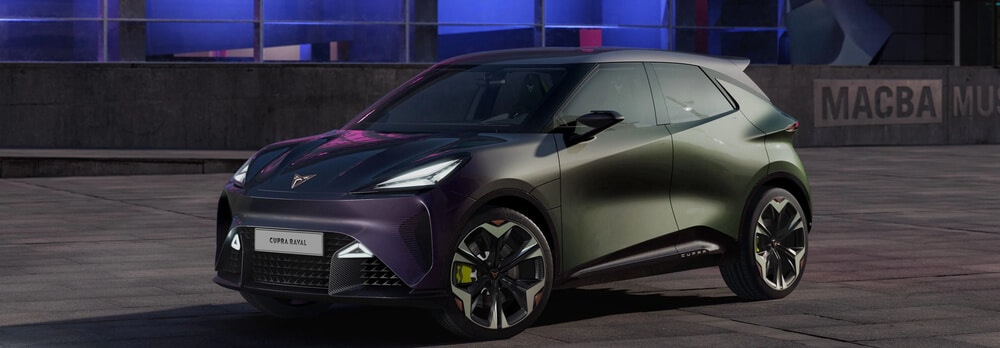 Imagen de un automóvil Cupra Raval estacionado en un entorno urbano moderno. El coche tiene un diseño futurista con líneas aerodinámicas y una pintura que cambia de color entre verde y púrpura según el ángulo de visión. Sus llantas son grandes y tienen un diseño geométrico distintivo. En la parte frontal, se observa el logotipo de Cupra y una placa con el nombre \"CUPRA RAVAL\". Al fondo, un edificio con ventanas iluminadas en tonos azules y morados, junto a una pared con el texto \"MACBA MUS\" visible, enmarca la escena.