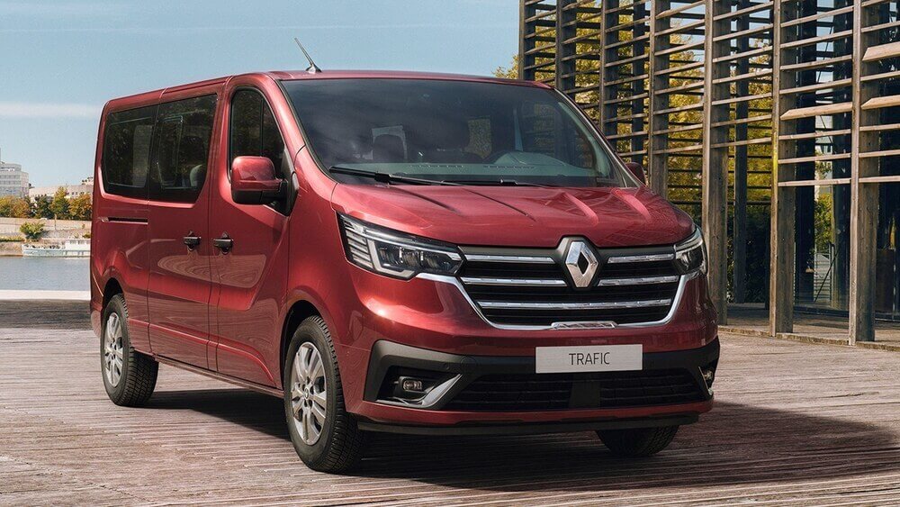 Furgoneta Renault Master en color rojo, con diseño robusto y aerodinámico. Se distingue por su gran capacidad de carga y sus puertas traseras de doble apertura. En la parte frontal, se observa el icónico logotipo de Renault sobre la parrilla negra, acompañado de faros de diseño moderno. La furgoneta está estacionada en una zona industrial con edificios comerciales en el fondo.