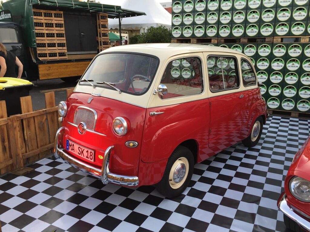 Un Fiat 600 Multipla clásico, de colores rojo y crema, está estacionado sobre un suelo ajedrezado en blanco y negro. A su alrededor hay un cerco de madera en un lado y pilas de barriles de cerveza Echbaum en otro. En el fondo, se observa un camión verde con cajas de madera y una persona de pie cerca de él. El Fiat 600 Multipla destaca por su diseño icónico y su importancia histórica como uno de los primeros vehículos multipropósito. La matrícula del coche es 'MA SK 18'.