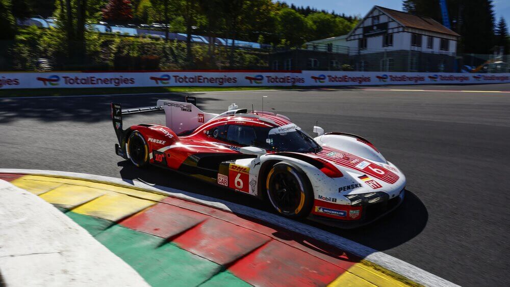 Un coche de carreras Porsche con el número 6 toma una curva en el circuito de Spa-Francorchamps. Su diseño aerodinámico, con colores rojo, blanco y negro, resalta mientras los logotipos de patrocinadores como Penske, Mobil 1 y Michelin adornan su carrocería. En el fondo, se pueden ver árboles, una casa y vallas publicitarias de TotalEnergies, añadiendo contexto a la escena de alta velocidad.