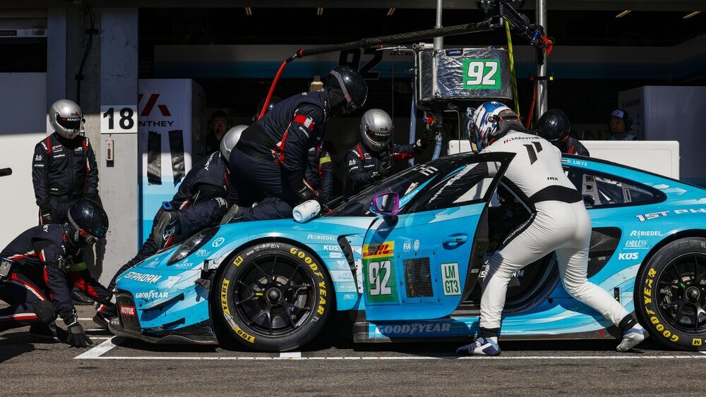 "Un Porsche 911 GT3 azul claro con los logotipos de Motul y Goodyear lidera una carrera en el circuito de Spa-Francorchamps. Detrás de él, varios autos de competición de diferentes colores y marcas siguen la acción de cerca. La escena captura la velocidad y la emoción de la carrera mientras los vehículos atraviesan una curva con bordes pintados en verde, amarillo y rojo, típicos de este icónico circuito.