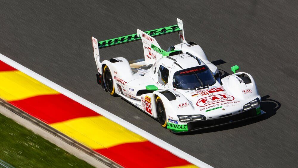 Un Porsche de carreras de color blanco con detalles en rojo y el número 6 avanza a toda velocidad por el circuito de Spa-Francorchamps. Su diseño aerodinámico resalta mientras las marcas 'Penske' y 'HY' adornan la parte delantera. En el fondo, otro coche amarillo sigue la acción, acentuando la sensación de velocidad y competencia.