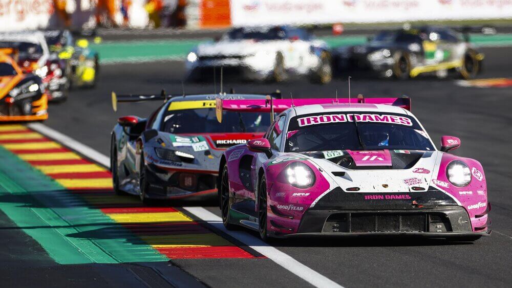"Un Porsche 911 GT3 R con los colores rosa y blanco del equipo 'Iron Dames' lidera la carrera en el circuito de Spa-Francorchamps. Detrás de él, otro coche de carreras con colores oscuros y detalles en amarillo sigue de cerca. La imagen captura la intensidad y la velocidad de la competencia mientras los autos toman una curva del circuito.