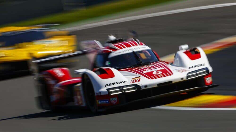 Un coche de carreras Porsche blanco con detalles en rojo y el número 6 avanza a gran velocidad por el circuito de Spa-Francorchamps. En la parte delantera, se pueden leer las palabras 'Penske' y 'HY'. Su movimiento se destaca por el desenfoque del fondo y las ruedas. Detrás del Porsche, otro coche de carreras amarillo también está en acción, capturando la velocidad y emoción de la competencia en este icónico circuito.