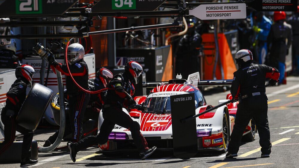 Un coche de carreras Porsche con el número 6 está en su parada en boxes durante una carrera en el circuito de Spa-Francorchamps. Un equipo de mecánicos, vestidos con monos rojos y negros, trabaja rápidamente para realizar tareas como cambiar los neumáticos y repostar combustible. La escena captura la acción intensa y la precisión necesarias en un pit stop para mantener la competitividad en la carrera.