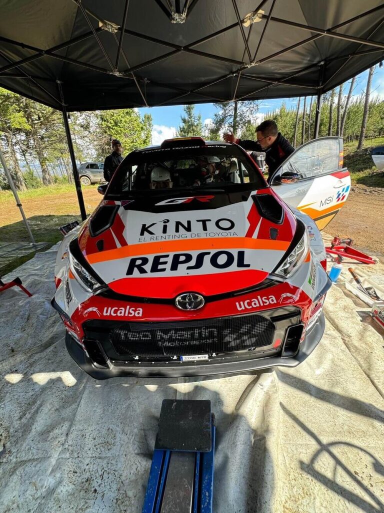 Coche de rally Toyota GR estacionado bajo una carpa en un entorno exterior. En el vehículo se observan los logotipos de KINTO, REPSOL y ucalsa como patrocinadores. A su lado derecho, una persona trabaja en la preparación o revisión del coche antes de la competición.