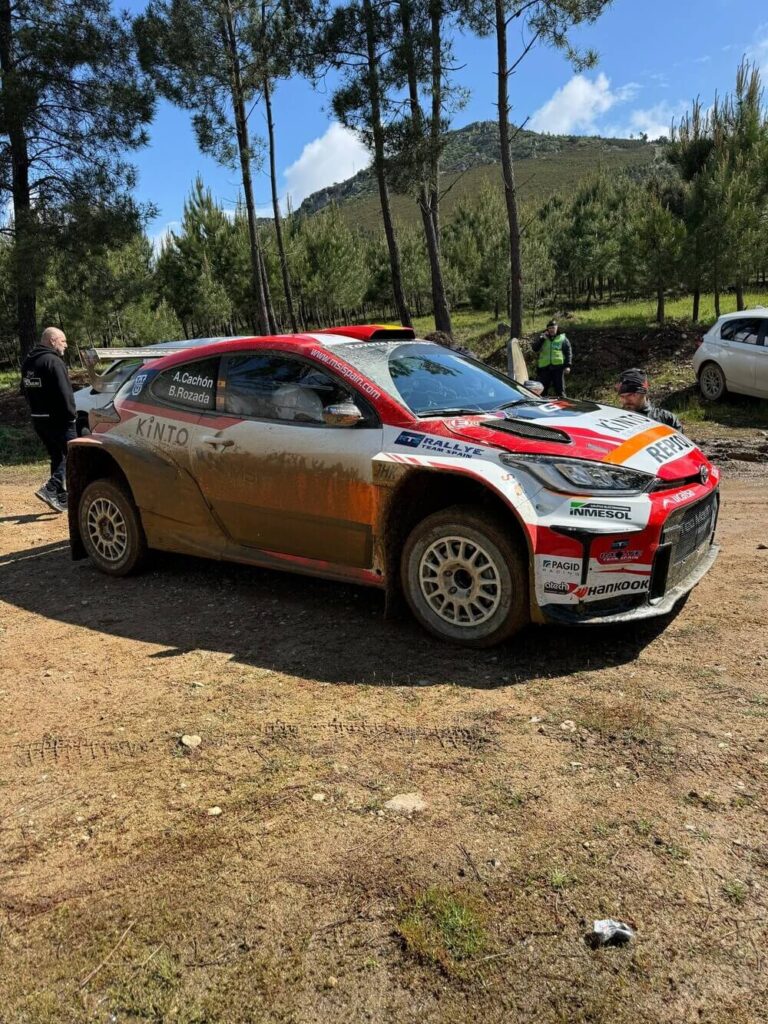 Coche de rally Toyota GR Yaris, cubierto de barro, con los nombres de los pilotos A. Cachón y B. Rozada en la ventana lateral. Lleva logotipos de patrocinadores como KINTO, Rallye Team Spain, Repsol y Hankook. Está en un entorno natural con árboles y montañas al fondo, rodeado por algunas personas, posiblemente miembros del equipo o espectadores.