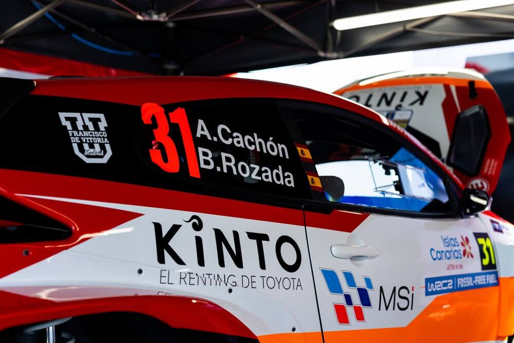 "Coche de carreras Toyota número 31 en un entorno de taller. En la ventana lateral, los nombres de los pilotos 'A. Cachón' y 'B. Rozada' junto a banderas de España. La carrocería muestra varios logotipos y patrocinadores, incluyendo 'Francisco de Vitoria Madrid', 'KINTO EL RENTING DE TOYOTA', 'MSi', 'Islas Canarias', y 'WRC2 FOSSIL-FREE FUEL'. Luz brillante en el techo.