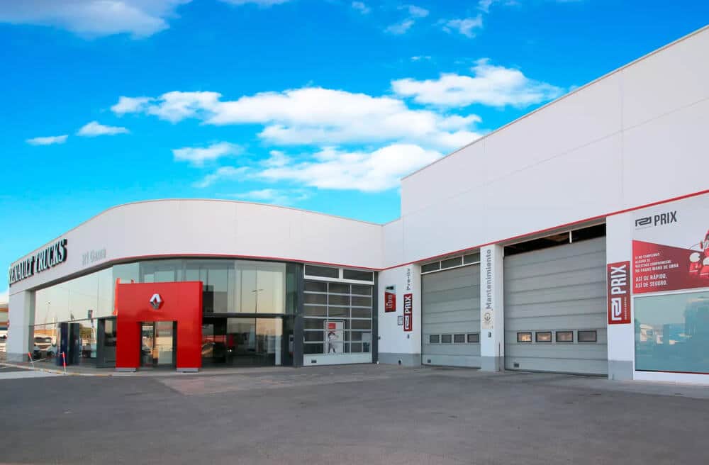 Taller de camiones Renault Trucks de la empresa R1Gama. En el edificio principal se observa el logotipo de Renault y el nombre 'RENAULT TRUCKS' en letras grandes y luminosas. Hay varias bahías de servicio donde se están realizando trabajos de mantenimiento en camiones de gran tamaño. En la parte exterior, se aprecia un camión estacionado, listo para ser atendido. La zona está equipada con herramientas y señalización propia de un centro de servicio especializado.