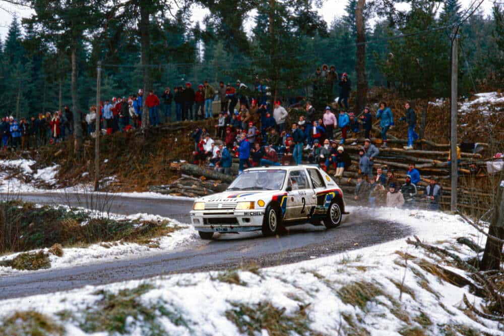 Un Peugeot 205 T16 de rally, conducido por Ari Vatanen, avanza por una carretera nevada y mojada, tomando una curva cerrada. El coche levanta agua y nieve mientras maniobra, mostrando su potencia y precisión. A lo lejos, una multitud de espectadores observa desde una zona elevada, protegida por una barrera de troncos. El entorno está rodeado de árboles y nieve, destacando las duras condiciones del rally en un paisaje invernal.