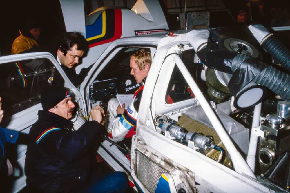 Ari Vatanen dentro de su Peugeot 205 T16 de rally, sentado en el asiento del conductor mientras su equipo técnico revisa el vehículo. El coche de competición tiene estructura de seguridad visible y componentes mecánicos expuestos, mostrando la preparación antes de una carrera.