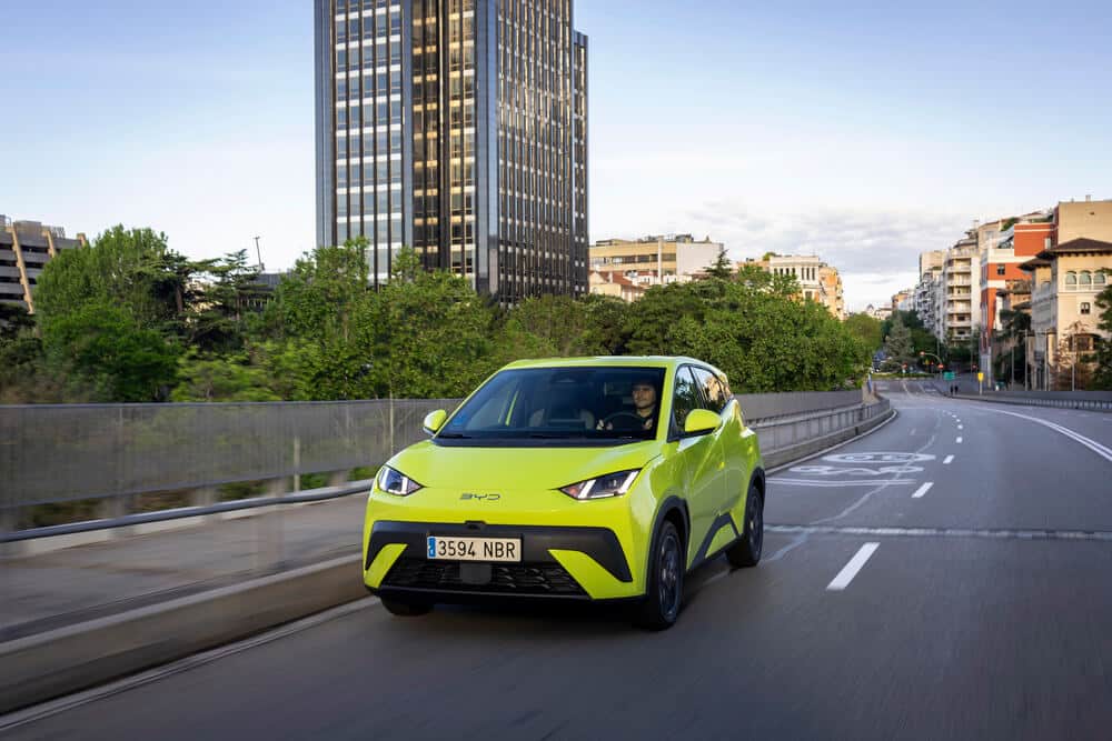 Un coche BYD de color verde brillante circula por una carretera urbana. El vehículo tiene un diseño moderno y aerodinámico, con luces delanteras angulares y una matrícula que dice '3594 NBR'. Al fondo, se pueden ver edificios altos y árboles, lo que sugiere que el coche está en una ciudad. La imagen resalta la integración de vehículos eléctricos en entornos urbanos.
