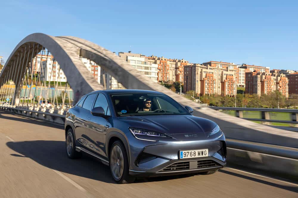 Un coche BYD gris oscuro cruza un puente moderno con un arco grande y cables de soporte. El vehículo tiene matrícula 9768 MWG y se ve una persona conduciendo. En el fondo, hay edificios residenciales de varios pisos y un cielo despejado.