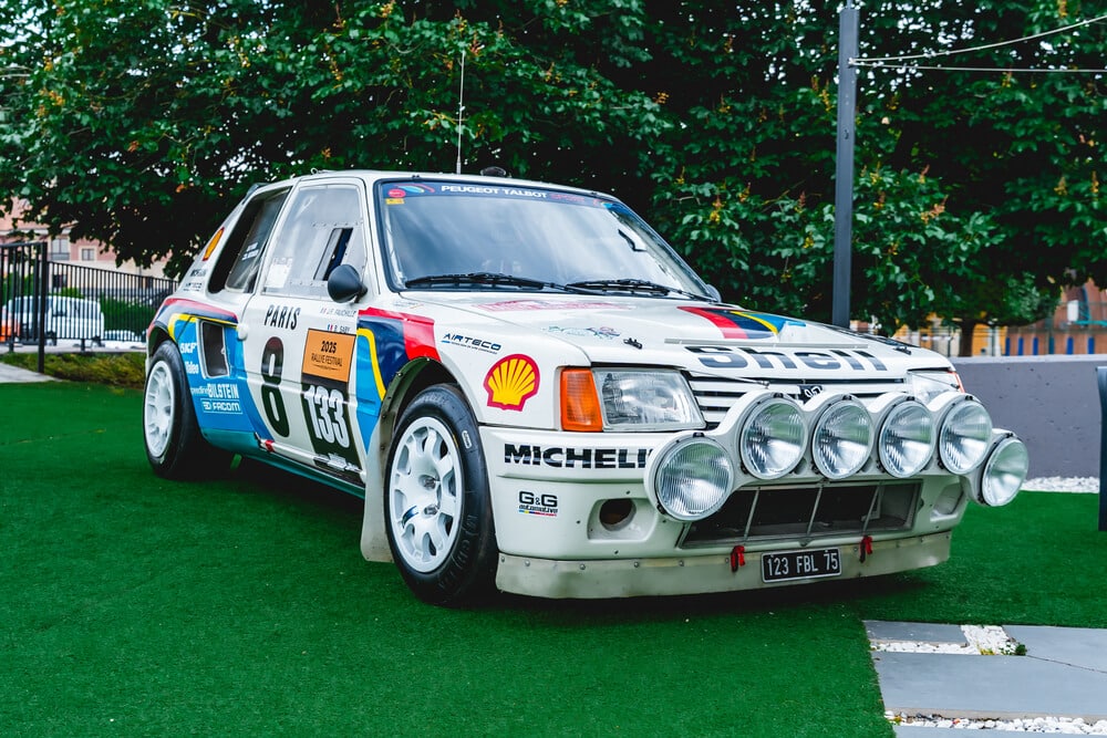 Un Peugeot 205 Turbo 16 de rally, conducido por Ari Vatanen, decorado con los colores y logotipos de patrocinadores como Shell y Michelin. En la puerta del coche se ven los números 8 y 1333, y en la parte frontal hay seis luces adicionales para mejorar la visibilidad nocturna. La matrícula es '123 FBL 75'. El coche está estacionado sobre césped artificial, con árboles y edificios en el fondo.