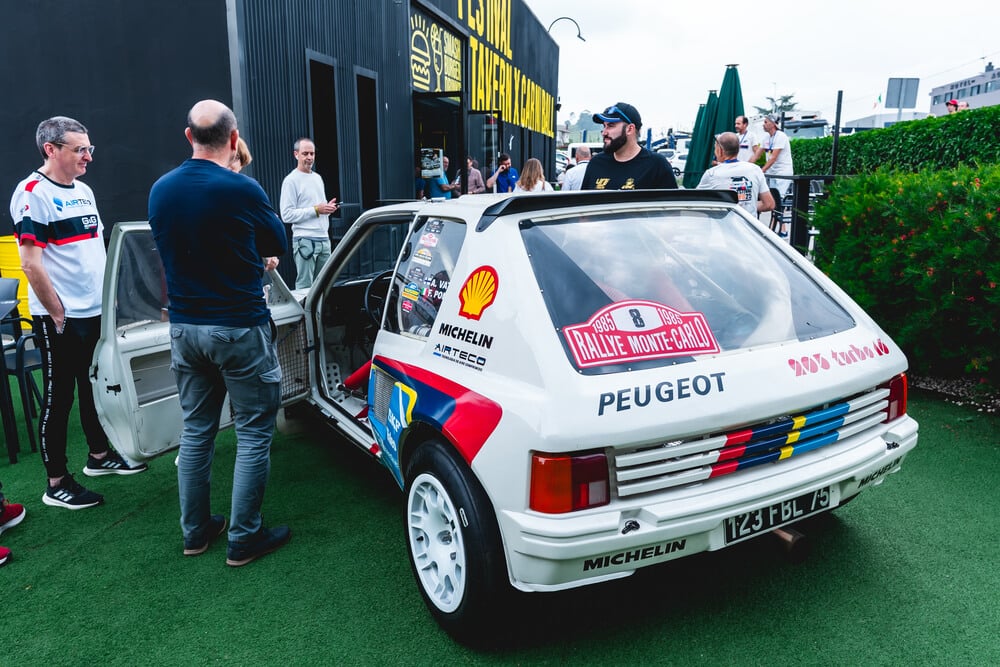 Imagen de un Peugeot 205 Turbo 16 con la icónica decoración del Rallye Monte-Carlo de 1985, conducido por Ari Vatanen. El vehículo está estacionado en un área al aire libre, rodeado de varias personas que observan su interior. La carrocería luce los logotipos de Michelin, Shell y Airteco, y el número 8 del rally. La matrícula es '123 FBL 75'. Un coche histórico que marcó una era en el mundo del rally.