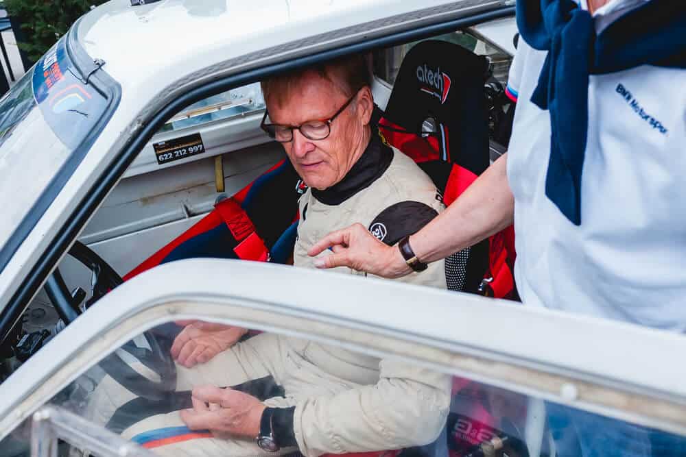 El famoso piloto de rally Ari Vatanen está sentado en el asiento del conductor de un automóvil de carreras. Lleva puesto un traje de carreras y está acompañado por otra persona vestida con una camisa blanca y un pañuelo azul, quien tiene una mano sobre su hombro. El automóvil tiene un interior simplificado y está equipado con un asiento de carreras con la marca "atech".