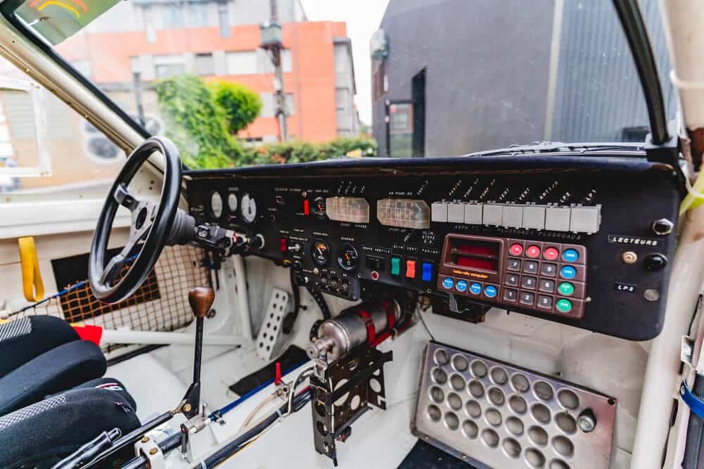 El interior de un coche de rally, específicamente un Peugeot 205 T16 de Ari Vatanen. La imagen muestra el tablero de instrumentos del coche, que incluye un volante negro, una palanca de cambios con pomo de madera y una consola llena de interruptores, botones y medidores. Hay una serie de indicadores analógicos y digitales, así como un teclado numérico en la parte derecha del tablero. El coche está equipado con asientos de competición y una jaula antivuelco.