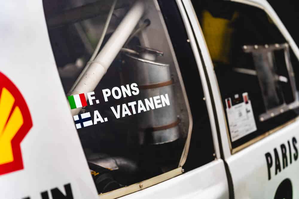 Primer plano de un Peugeot T16 de competición. En la ventana lateral se leen los nombres 'F. Pons' con bandera italiana y 'A. Vatanen' con bandera finlandesa. En la carrocería hay un logotipo de Shell. A través de la ventana se observan componentes mecánicos, como una barra de refuerzo y un tanque metálico, que resaltan la ingeniería del vehículo.