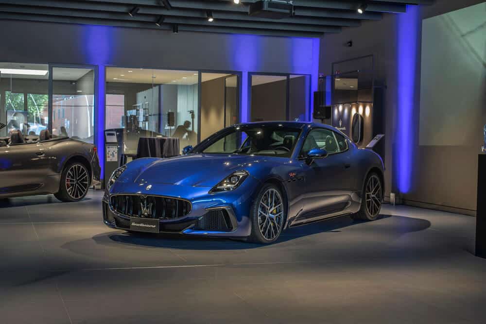 Un Maserati GranTurismo azul exhibido en un showroom moderno con iluminación azul. Se aprecia otro Maserati parcialmente visible en el fondo.