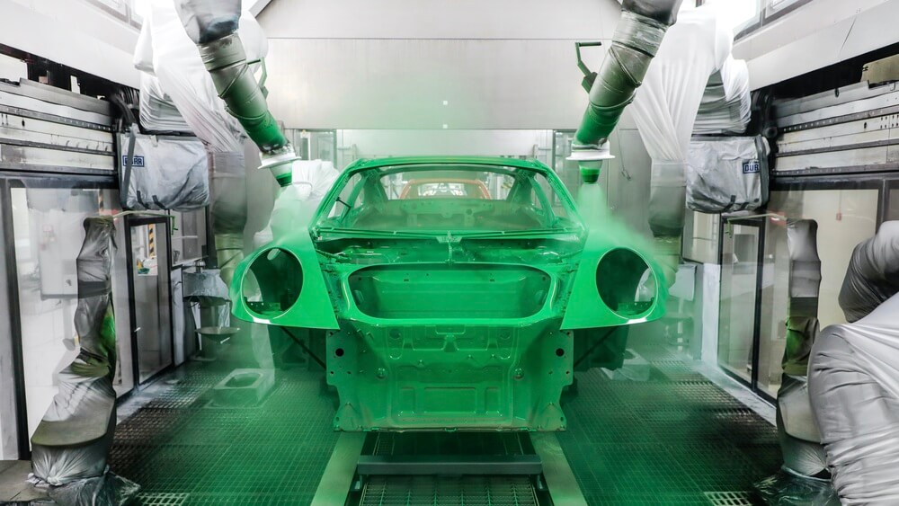 Un automóvil Porsche en una línea de producción de pintura. Varios brazos robóticos, cubiertos con fundas protectoras, rocían pintura verde sobre la carrocería del vehículo dentro de una cabina automatizada. El proceso de aplicación es preciso y meticuloso, destacando la tecnología avanzada utilizada en la fabricación de automóviles para garantizar un acabado impecable.