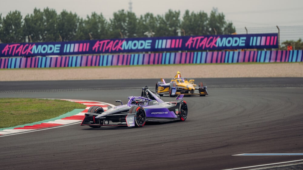 Dos coches de Fórmula E compiten en la pista, tomando una curva a alta velocidad. En primer plano, un Porsche negro y púrpura lidera, seguido de un vehículo amarillo. Detrás de ellos, una valla publicitaria con el texto 'ATTACK MODE' resalta en colores vibrantes, enmarcando la acción de la carrera.
