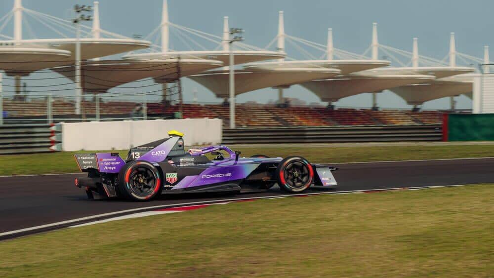 "Coche de carreras de Fórmula E número 13 en una curva de la pista, con colores morado y negro y logotipos de patrocinadores como TAG Heuer, Porsche, Cato, Ansys y Hankook. En primer plano, césped junto a la pista; al fondo, gradas y estructuras blancas con forma de sombrilla cubriendo parte del público.