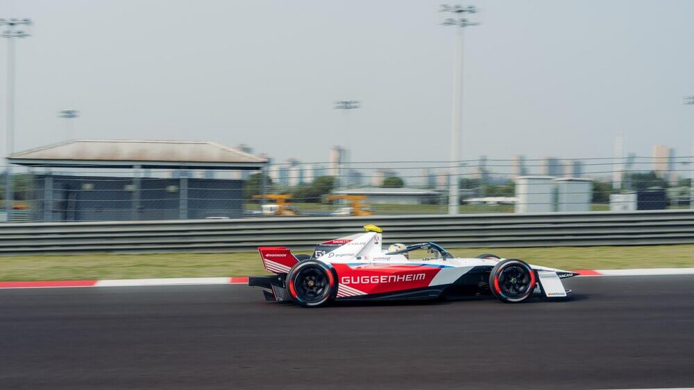 "Imagen de un coche de carreras de Fórmula E en movimiento sobre una pista. El vehículo es eléctrico, con un diseño aerodinámico y colores blanco, rojo y negro. En el lateral se puede leer 'GUGGENHEIM'. La pista tiene bordes pintados de rojo y blanco. De fondo, se ven estructuras y edificios bajo luces altas. La imagen captura la velocidad y modernidad de la Fórmula E.