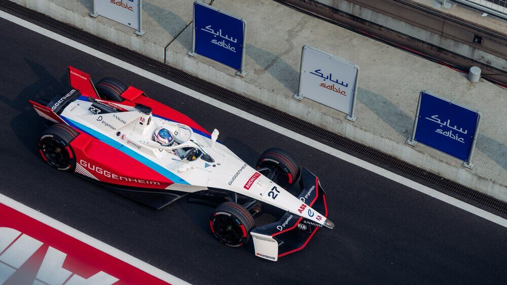 Imagen de un coche de Fórmula E en la pista. El coche es de color blanco con detalles en rojo y azul, y tiene el número 27 en la parte delantera. En el lateral del coche se puede leer "GUGGENHEIM". El piloto está dentro del coche, con casco azul y blanco. Al fondo, se ven tres carteles con la palabra "sabic" escrita en ellos.