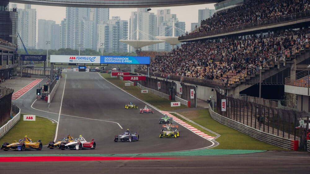 Varios coches de Fórmula E toman una curva en el circuito de Shanghái, con espectadores llenando las gradas a la derecha. Al fondo, el paisaje urbano de Shanghái con edificios altos. En la parte superior, un cartel indica 'ABB Formula E FIA World Championship 2025 Hankook Shanghai E-Prix'.
