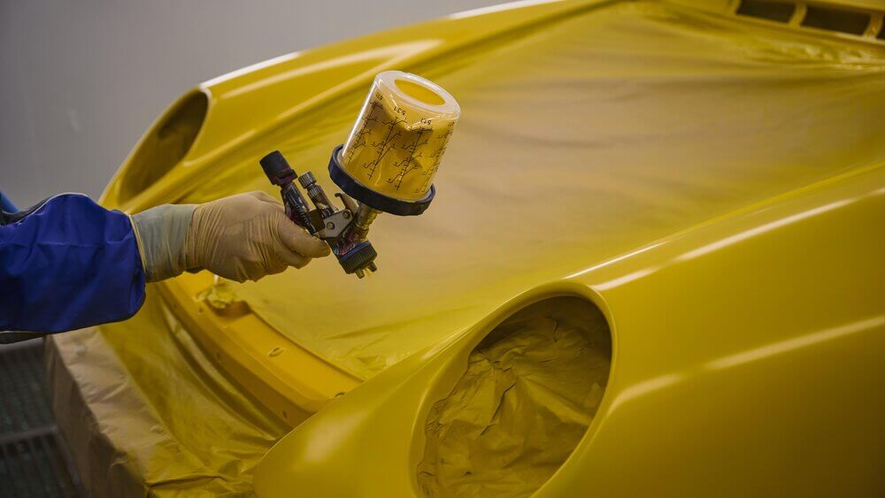 Una persona está pintando un Porsche amarillo con una pistola de pintura. La parte delantera del coche está cubierta con papel para proteger las áreas que no deben ser pintadas. La imagen captura el proceso de aplicación de la pintura, destacando una etapa clave en la restauración o personalización del vehículo.