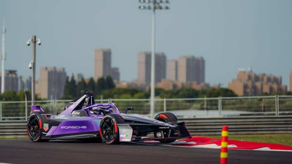 Imagen de un coche de carreras de Fórmula E con los colores morado y negro, con la marca Porsche visible en el lateral. El coche está en una pista de carreras, con edificios altos y árboles en el fondo. Hay una barrera de seguridad y un cono de señalización en primer plano.