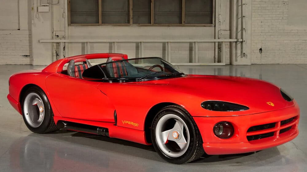 "Automóvil deportivo Dodge Viper RT/10 de color rojo, estacionado en un espacio interior con paredes blancas y grandes ventanas. El coche tiene un diseño aerodinámico con faros delanteros oscuros y alargados, llantas plateadas de cinco radios y el emblema 'Viper RT/10' en amarillo en la puerta del conductor.