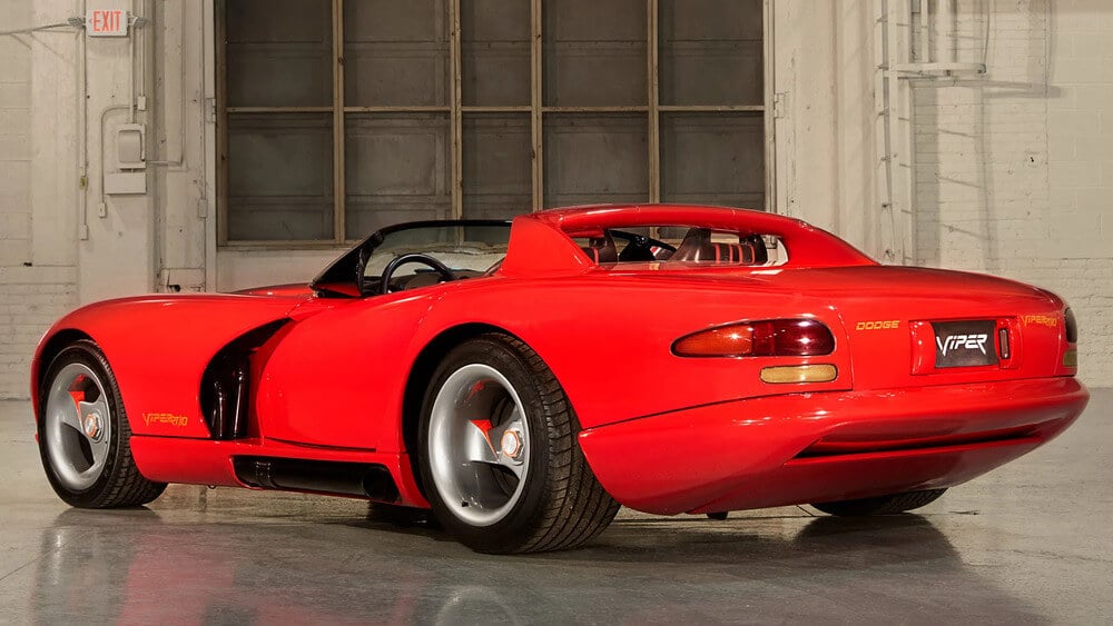 Automóvil deportivo Dodge Viper RT/10 de color rojo, estacionado en un espacio interior de estilo industrial con paredes blancas y grandes ventanas. El coche presenta un diseño aerodinámico clásico, con llantas plateadas, detalles en rojo y los emblemas 'Dodge' y 'Viper RT/10' visibles en la parte trasera.