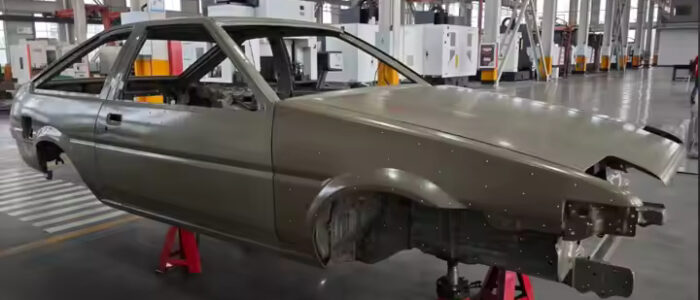 Carrocería de un Toyota AE86 en un taller industrial, montada sobre soportes rojos sin ruedas. La carrocería es de color plateado y está en proceso de restauración o ensamblaje, sin componentes externos como faros y parachoques. El fondo muestra un espacio limpio y bien iluminado con columnas estructurales y maquinaria al fondo.