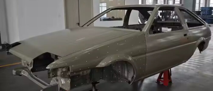 Carrocería de un Toyota AE86 en un taller industrial, montada sobre soportes rojos sin ruedas. La carrocería es de color plateado y está en proceso de restauración o ensamblaje, sin componentes externos como faros y parachoques. El fondo muestra un espacio limpio y bien iluminado con columnas estructurales y maquinaria al fondo.