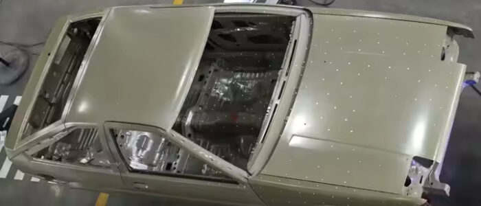 Vista superior de la carrocería de un Toyota AE86 sin paneles exteriores, mostrando los componentes internos y el chasis. El coche está situado en un entorno interior, probablemente un taller o garaje, con suelo de hormigón. Al lado del coche hay una hoja de papel o tablero blanco en el suelo.