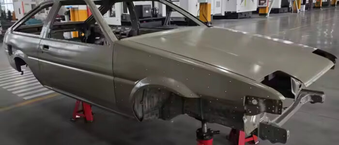 Carrocería de un Toyota AE86 soportada por soportes rojos en un entorno industrial o de fábrica. La carrocería está incompleta, sin ruedas, motor, componentes interiores ni otras partes esenciales, mostrando la estructura básica del vehículo en una etapa temprana de ensamblaje.