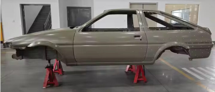 Carrocería de un Toyota AE86 soportada sobre cuatro soportes rojos en el interior de un edificio, probablemente un taller o garaje. El coche no tiene ruedas, motor ni otras partes externas; es solo el marco de metal desnudo.