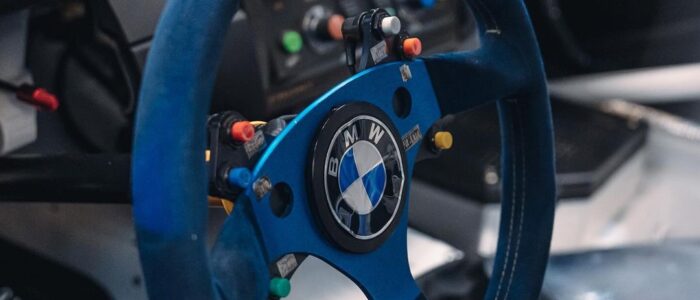 BMW M3 GTR Most Wanted (4) La imagen muestra el interior de un BMW M3 GTR, famoso por su aparición en el videojuego "Need for Speed: Most Wanted". El enfoque está en el volante y el tablero de instrumentos. El volante es azul con el logotipo de BMW en el centro y cuenta con múltiples botones y interruptores, probablemente para controlar varias funciones del coche. El tablero detrás del volante tiene varios botones e interruptores, indicando un alto nivel de personalización y funcionalidad típica en coches de carreras. Esta imagen es interesante y relevante porque destaca el equipo especializado y los controles utilizados en vehículos de carreras profesionales.