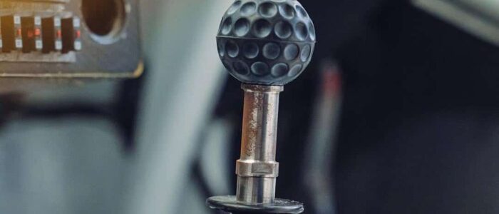 BMW M3 GTR Most Wanted (6) La imagen muestra el interior de un BMW M3 GTR, famoso por su aparición en el videojuego "Need for Speed: Most Wanted". El enfoque está en la palanca de cambios, que tiene un diseño único que se asemeja a una pelota de golf con una textura de hoyuelos. A la izquierda de la palanca de cambios, hay un panel con varios interruptores y una etiqueta que dice "FIRE". Esta imagen destaca un detalle interior personalizado o modificado, relevante para los entusiastas de los coches y aquellos interesados en modificaciones únicas.