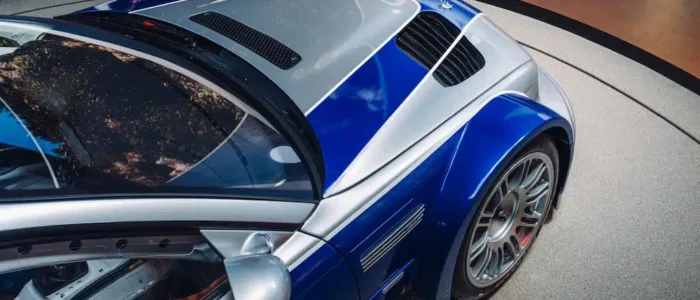 bmw-m3-gtr-nfs-hd-04 La imagen muestra un BMW M3 GTR, famoso por su aparición en el videojuego "Need for Speed: Most Wanted", en una pista de carreras. El coche tiene un diseño aerodinámico con un esquema de colores azul y blanco, y está equipado con un gran alerón trasero y llantas de aleación. La pista está rodeada de barreras de seguridad y el entorno sugiere un ambiente de competición. La imagen es interesante y relevante para los entusiastas del automovilismo y los fans del juego, destacando el coche en su elemento natural: la pista de carreras.