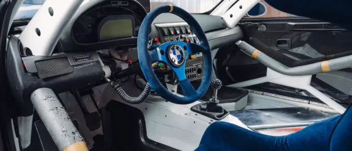 bmw-m3-gtr-nfs-hd-05 La imagen muestra el interior de un BMW M3 GTR, famoso por su aparición en el videojuego "Need for Speed: Most Wanted". El enfoque está en el volante azul con el logotipo de BMW en el centro, la palanca de cambios manual y varios interruptores y medidores en el tablero. El coche tiene una jaula antivuelco para mayor seguridad y los asientos están tapizados en tela azul. El interior está simplificado para reducir el peso, con componentes visibles de metal y fibra de carbono. Este diseño es interesante y relevante para aquellos interesados en las carreras automovilísticas, las modificaciones de coches o las características de seguridad en vehículos de alto rendimiento.