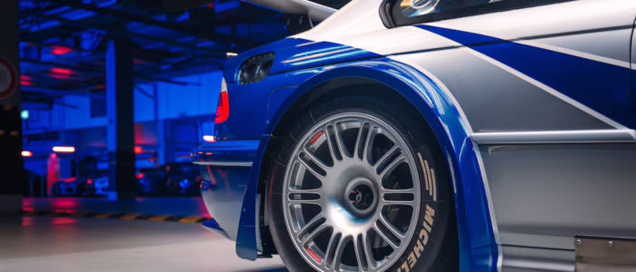 bmw-m3-gtr-nfs-hd-06 La imagen muestra la sección trasera de un BMW M3 GTR, famoso por su aparición en el videojuego "Need for Speed: Most Wanted". El enfoque está en la rueda trasera y parte de la carrocería del coche, que tiene un esquema de colores azul y plateado con detalles en blanco. El neumático lleva la marca "MICHELIN" y presenta un diseño detallado de llanta de aleación. El coche está en un garaje o taller bien iluminado con luces azules y rojas, añadiendo un efecto dramático. La presencia de un gran alerón trasero sugiere que es un vehículo de alto rendimiento, probablemente utilizado en carreras. La imagen es interesante porque destaca el diseño intrincado y la ingeniería de un coche de carreras, mostrando elementos como las características aerodinámicas y los neumáticos especializados.