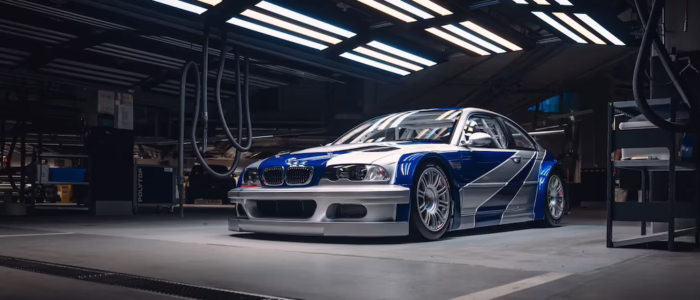 bmw-m3-gtr-nfs-hd-08 Ahora para esta del BMW M3 GTR de Need for Speed Most Wanted. No omitas esos detalles: La imagen muestra un BMW M3 GTR, un coche de carreras de alto rendimiento, estacionado en un garaje o taller bien iluminado. El coche presenta una distintiva librea azul y blanca con calcomanías de carreras, un kit de carrocería ancho y mejoras aerodinámicas, lo que lo hace visualmente llamativo. Este coche es particularmente interesante y relevante porque es famoso por el videojuego "Need for Speed: Most Wanted", donde juega un papel central. El entorno de la imagen, con su iluminación industrial y equipo, añade a la atmósfera de un entorno de carreras profesional.