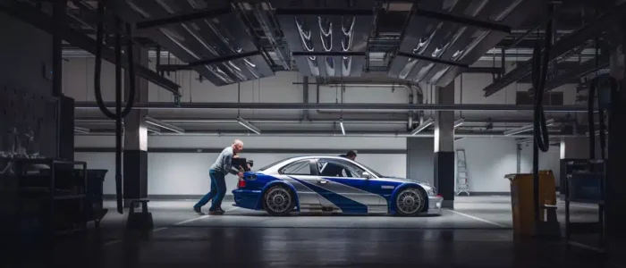 bmw-m3-gtr-nfs-hd-10 La imagen muestra un BMW M3 GTR, famoso por su aparición en el videojuego "Need for Speed: Most Wanted", siendo empujado por dos personas en un garaje bien iluminado. El coche tiene un diseño elegante con franjas de carreras y parece ser un vehículo de alto rendimiento. El garaje está equipado con varias herramientas y equipos, y el techo tiene múltiples luminarias. La escena captura un momento de preparación o mantenimiento del coche de carreras, destacando los esfuerzos detrás de escena en el automovilismo.