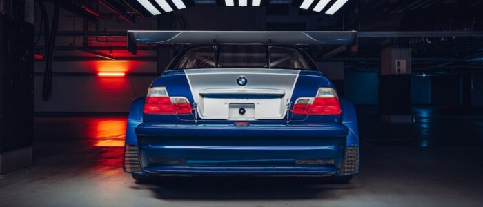 bmw-m3-gtr-nfs-hd-14 La imagen muestra la vista trasera de un BMW M3 GTR, famoso por su aparición en el videojuego "Need for Speed: Most Wanted". El coche está estacionado en un garaje con poca iluminación, lo que resalta su diseño aerodinámico y su gran alerón trasero. La iluminación crea un efecto dramático, destacando las líneas y características del coche, haciéndolo visualmente impactante y relevante para los entusiastas del automovilismo y los fans del juego.