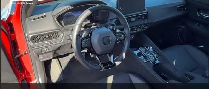 "El interior de un Honda Prelude 2026, específicamente el lado del conductor. Se pueden ver el volante, el tablero y la consola central. El volante tiene múltiples botones y hay una pantalla digital detrás de él. La consola central presenta varios controles y una pantalla grande. El texto en la imagen está en japonés y dice: 'タイプR譲りのフロントサスペンションを奢る,' que se traduce como 'Equipado con la suspensión delantera heredada del Type R.'
