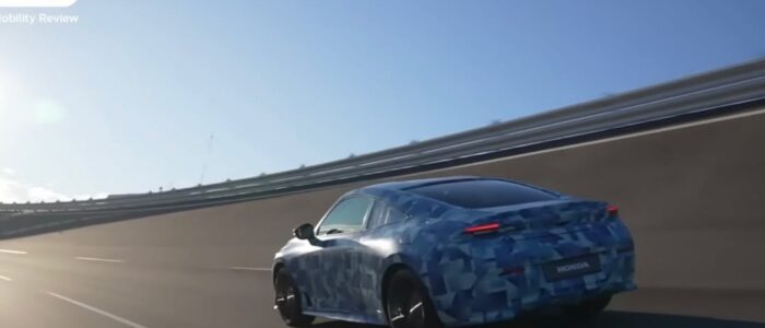 "Un Honda Prelude 2026 conduciendo por una autopista. El coche parece ser un prototipo o un vehículo de prueba, ya que está cubierto con un camuflaje azul. La autopista está vacía y el coche está conduciendo en una sección curva de la carretera. El sol brilla intensamente, creando sombras y un efecto de destello de lente. La imagen probablemente se tomó de un video promocional o de revisión, como sugiere el texto 'LIVE NOW' y 'Mobility Review' en la esquina superior izquierda.