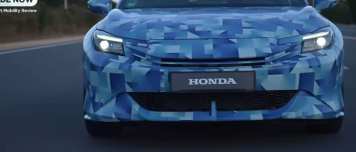 "Vista frontal de un coche Honda Prelude 2026 cubierto con un patrón de camuflaje azul, utilizado típicamente para ocultar los detalles de diseño de un nuevo modelo o prototipo durante las pruebas. Los faros están encendidos y el logotipo de Honda se muestra prominentemente en la parrilla frontal. El fondo muestra una carretera borrosa y algunos árboles, indicando que el coche está en movimiento.